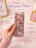 A hand holds a clear plastic package of 'Distant Mountain Flower' flower stickers. The package contains 15 pink and purple-toned flower stickers.